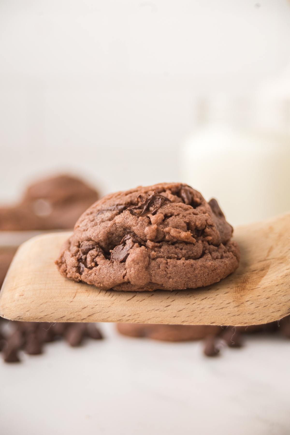 A chocolate cookie on a wooden spatula, with blurred cookies and milk in the background.