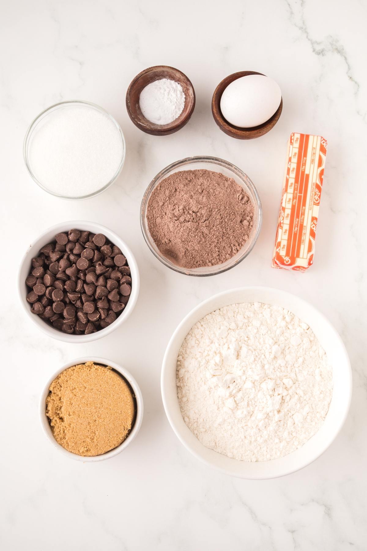 Baking ingredients on a white surface: flour, egg, butter, sugars, cocoa, chocolate chips, salt, and baking soda.