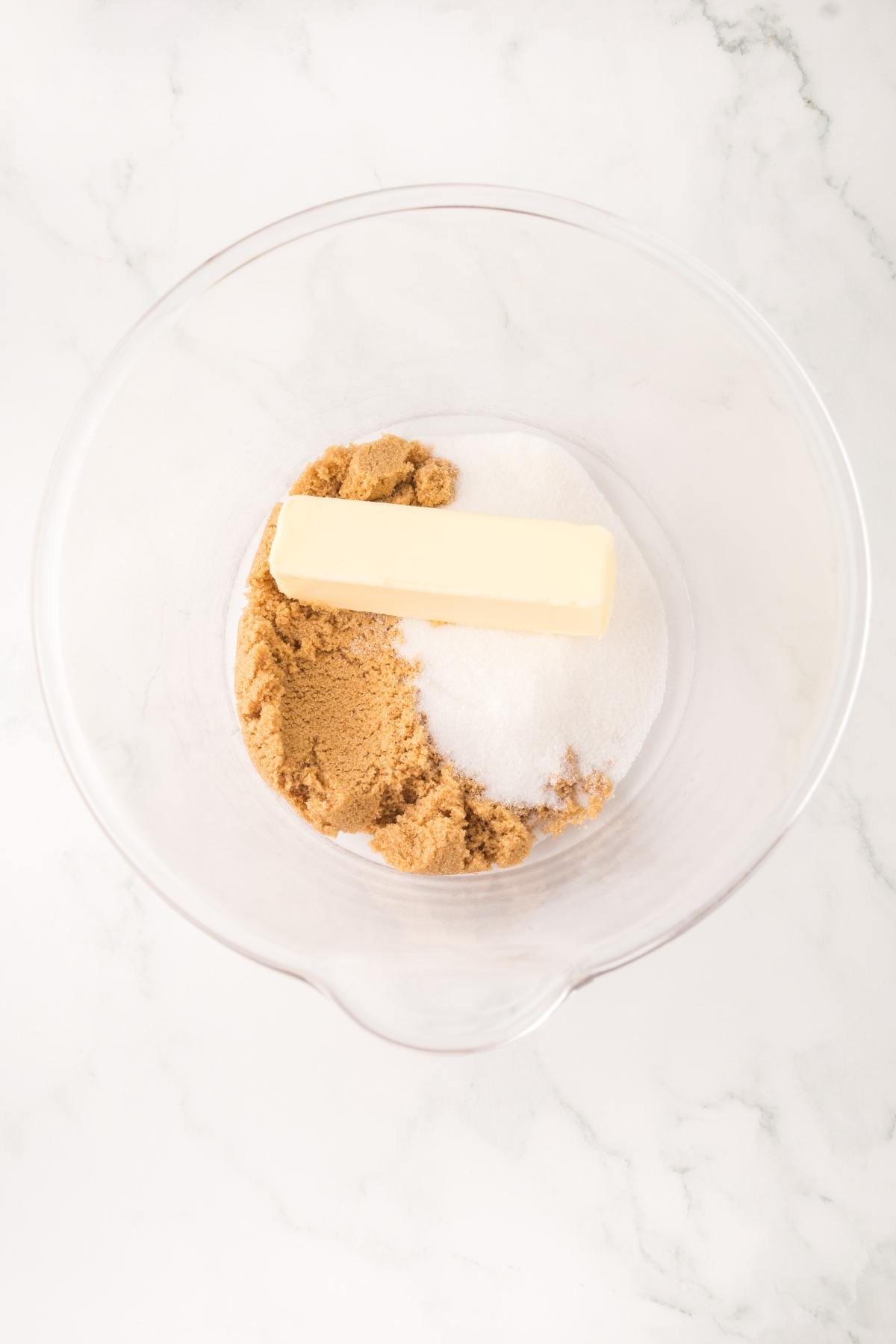 A glass bowl with a stick of butter, brown sugar, and white sugar on a white marble surface.