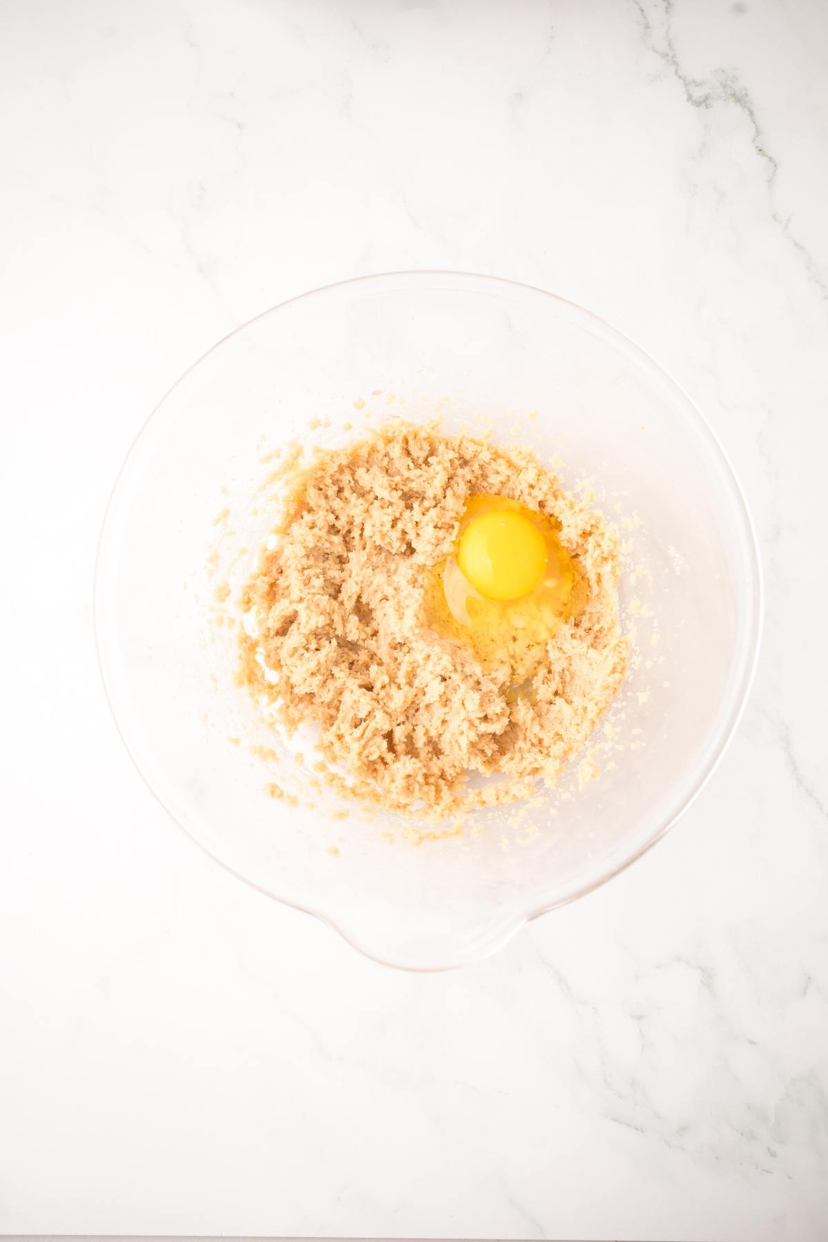 A clear bowl with creamed sugar and butter, and a cracked egg on top, on a white marble surface.