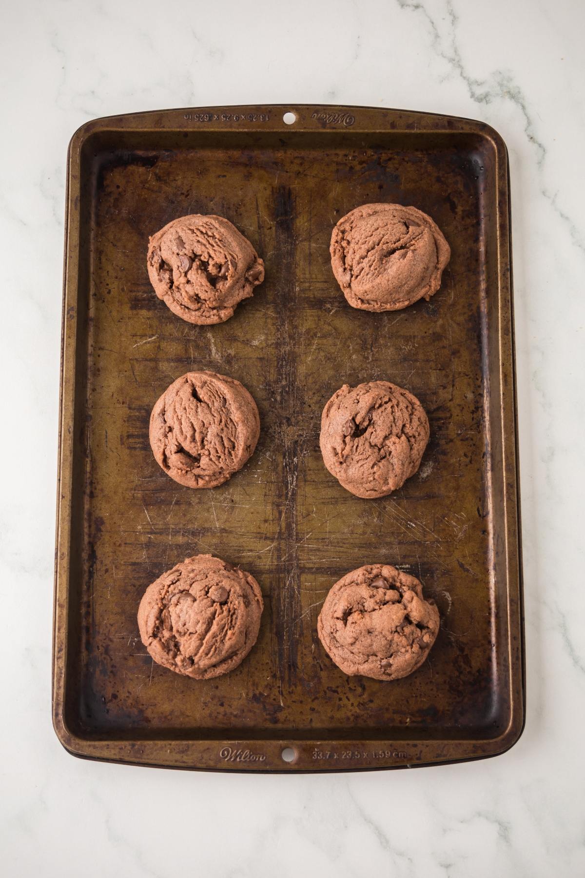 Six chocolate cookies on a worn baking sheet, placed on a white marble surface.