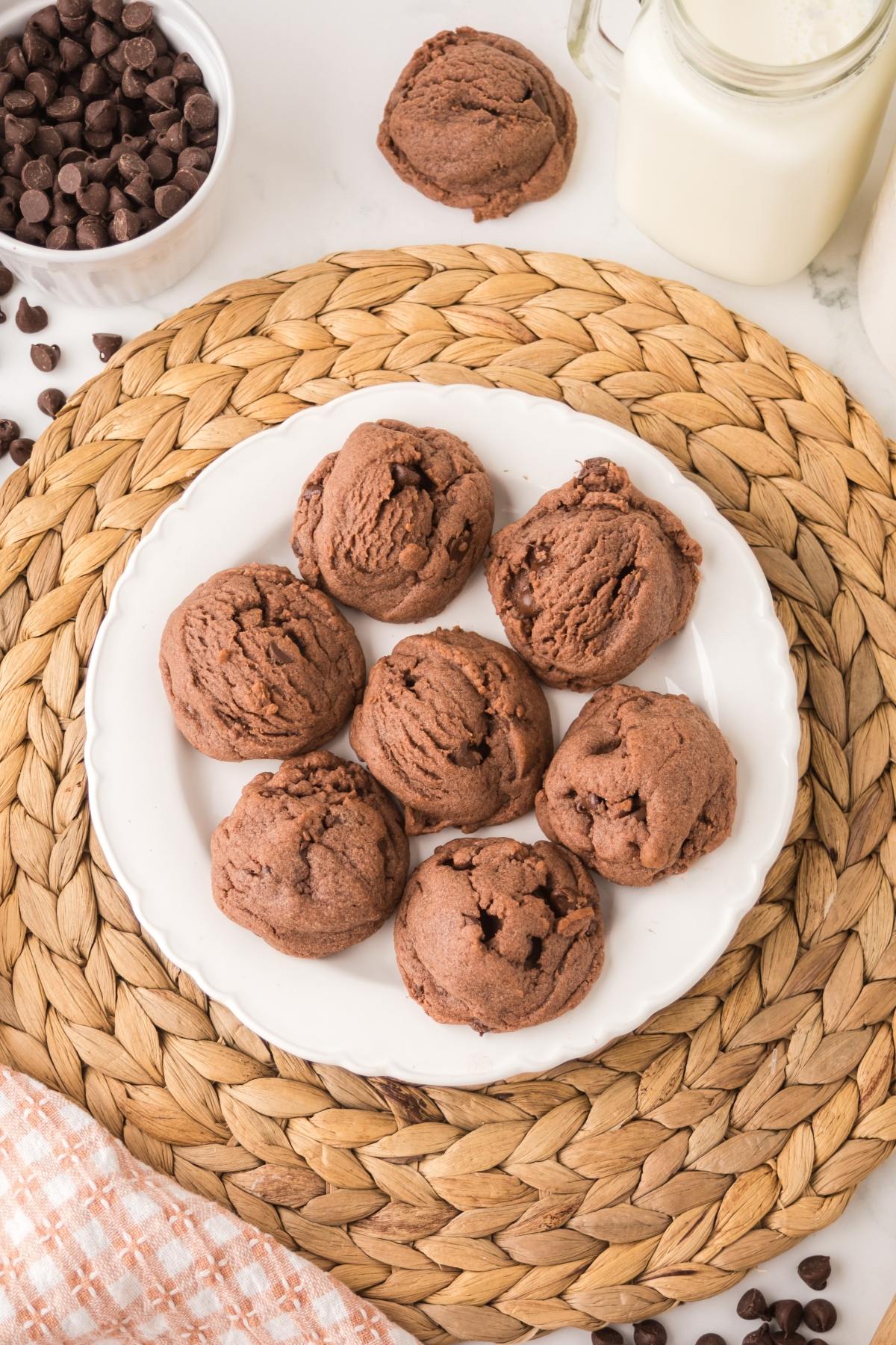 A white plate with seven chocolate cookies on a woven placemat, with milk and chocolate chips nearby.