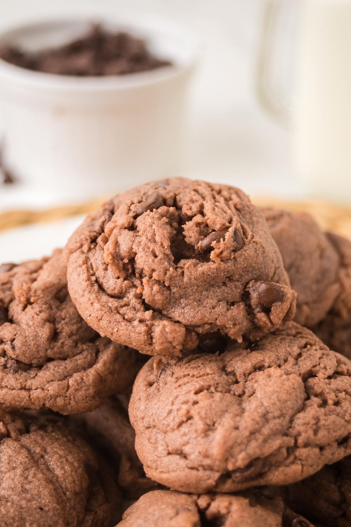 A close-up of a stack of soft, chocolate cookies with a blurred bowl and glass in the background.