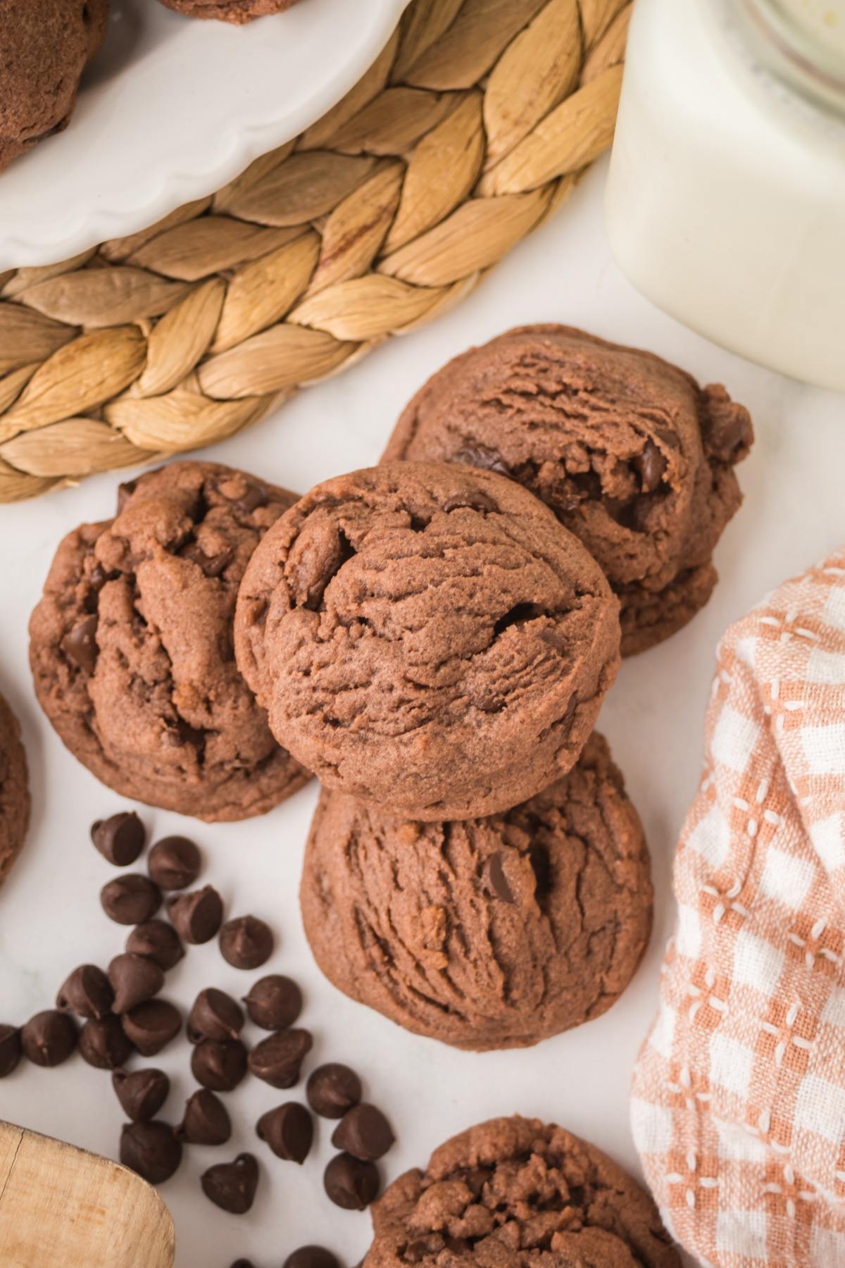 Three chocolate cookies stacked on a white surface near chocolate chips, a napkin, and a glass of milk.