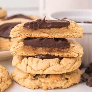 A stack of cookies topped with chocolate frosting, surrounded by chocolate chips, reminiscent of classic Chocolate Thumbprint Cookies.