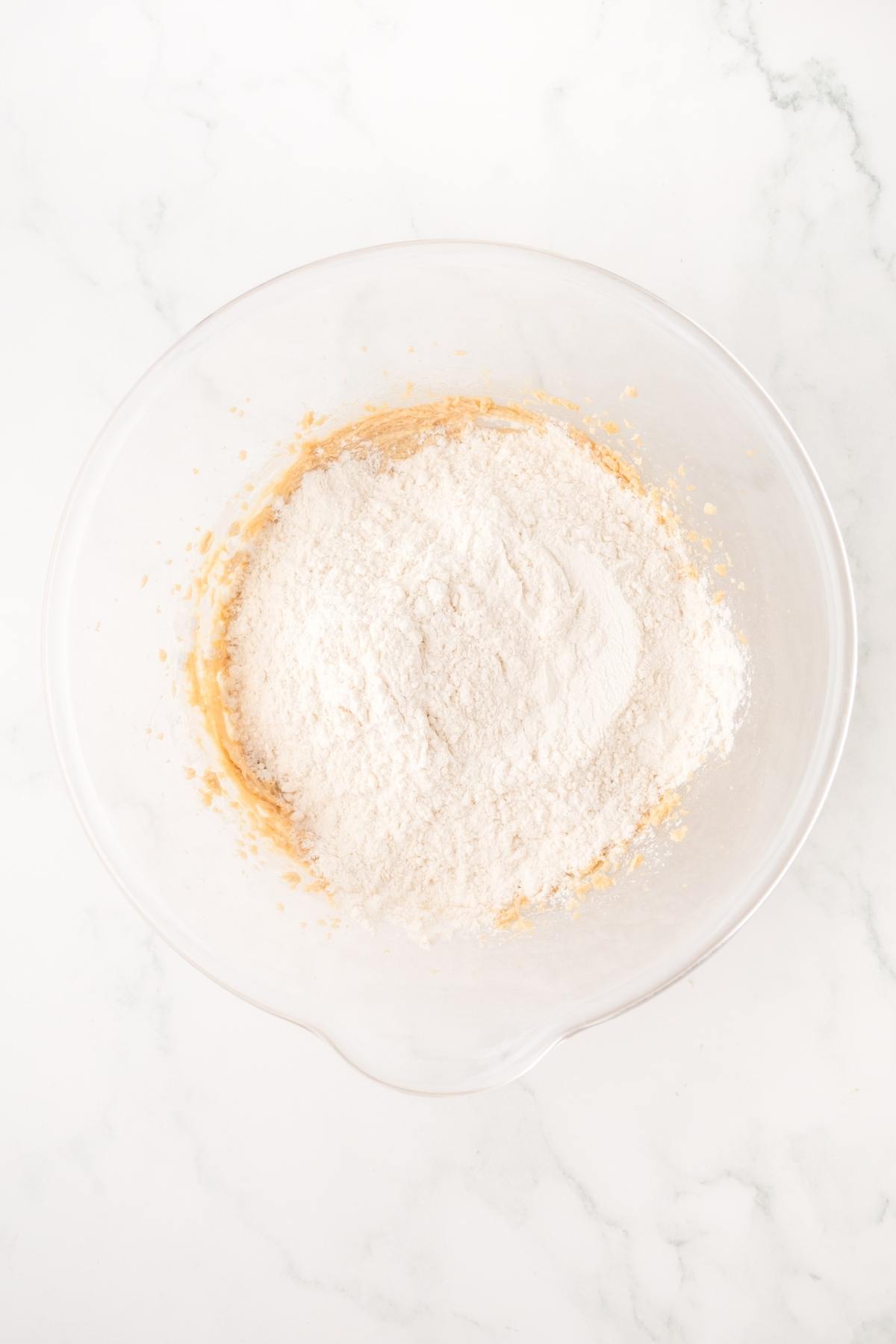 Clear mixing bowl with flour on top of dough mixture, on a white marble surface.