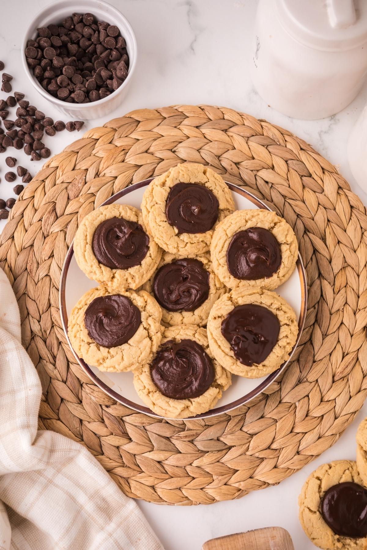 A plate of cookies with chocolate centers on a woven placemat, surrounded by chocolate chips and a cloth.