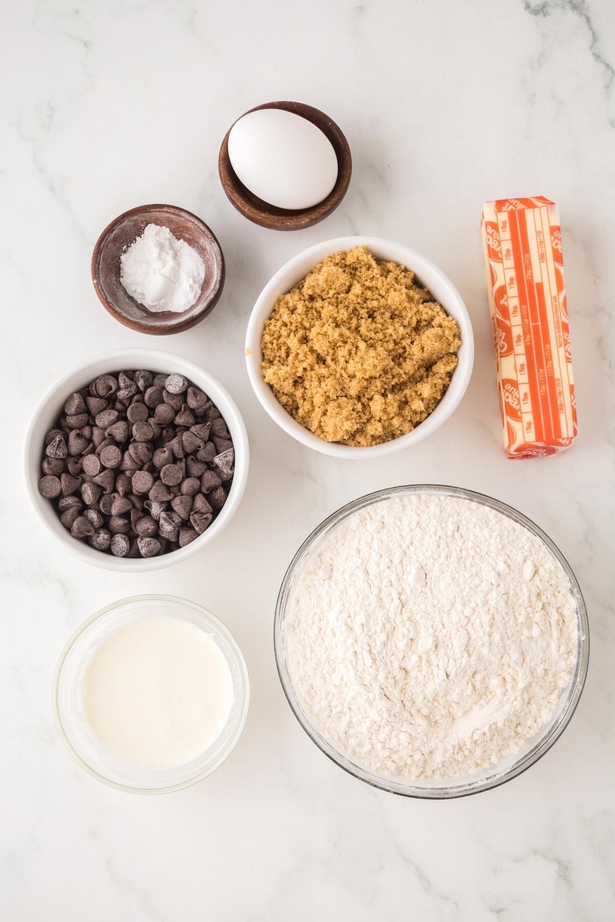 Baking ingredients on a white surface: egg, butter, brown sugar, flour, chocolate chips, milk, and baking soda.