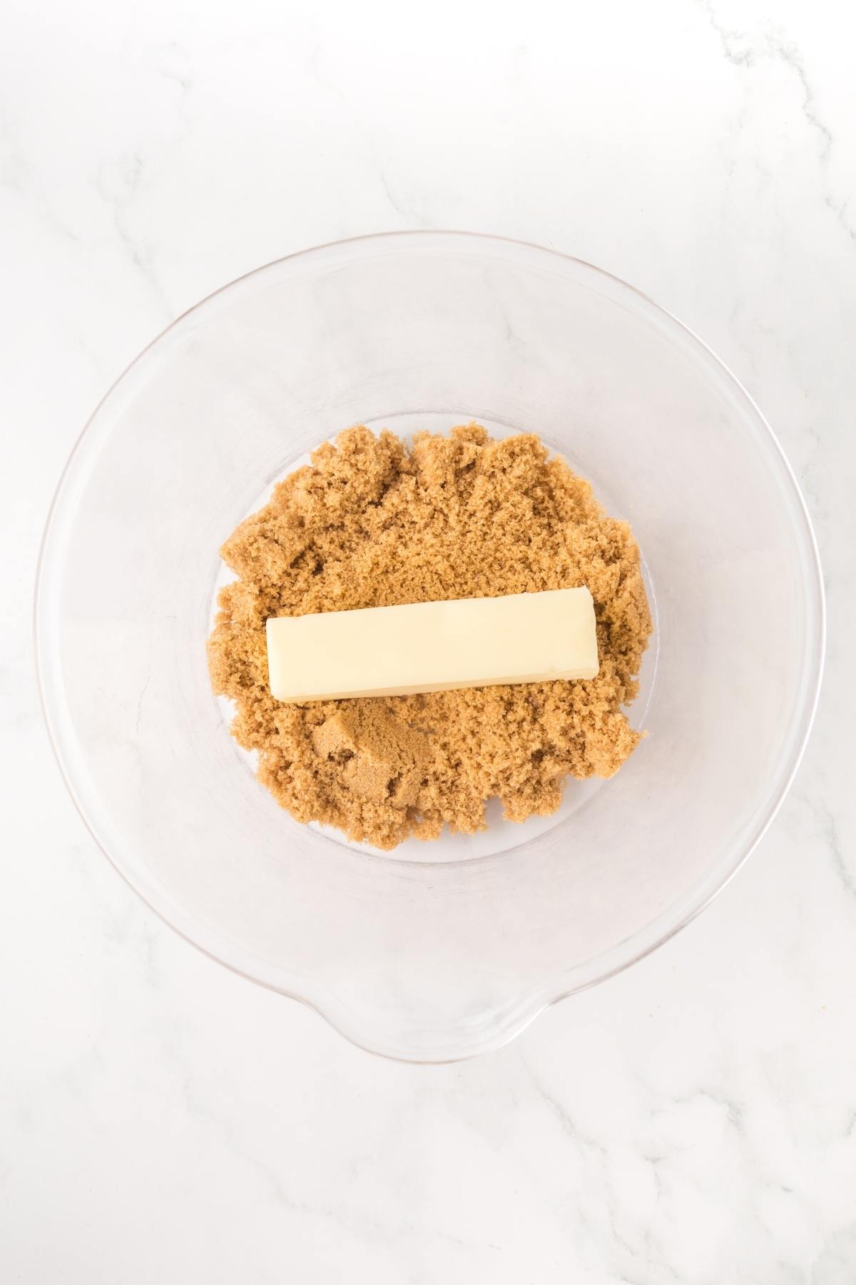 A stick of butter on top of brown sugar in a clear glass mixing bowl.