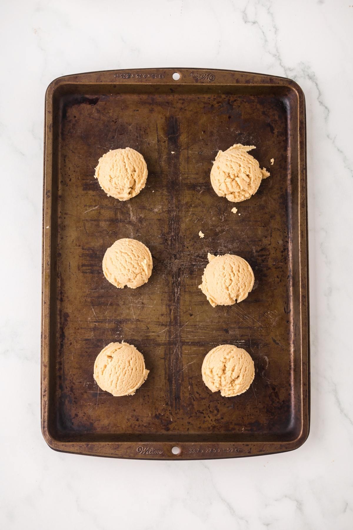Six scoops of cookie dough on a worn baking tray, placed on a white marble surface.
