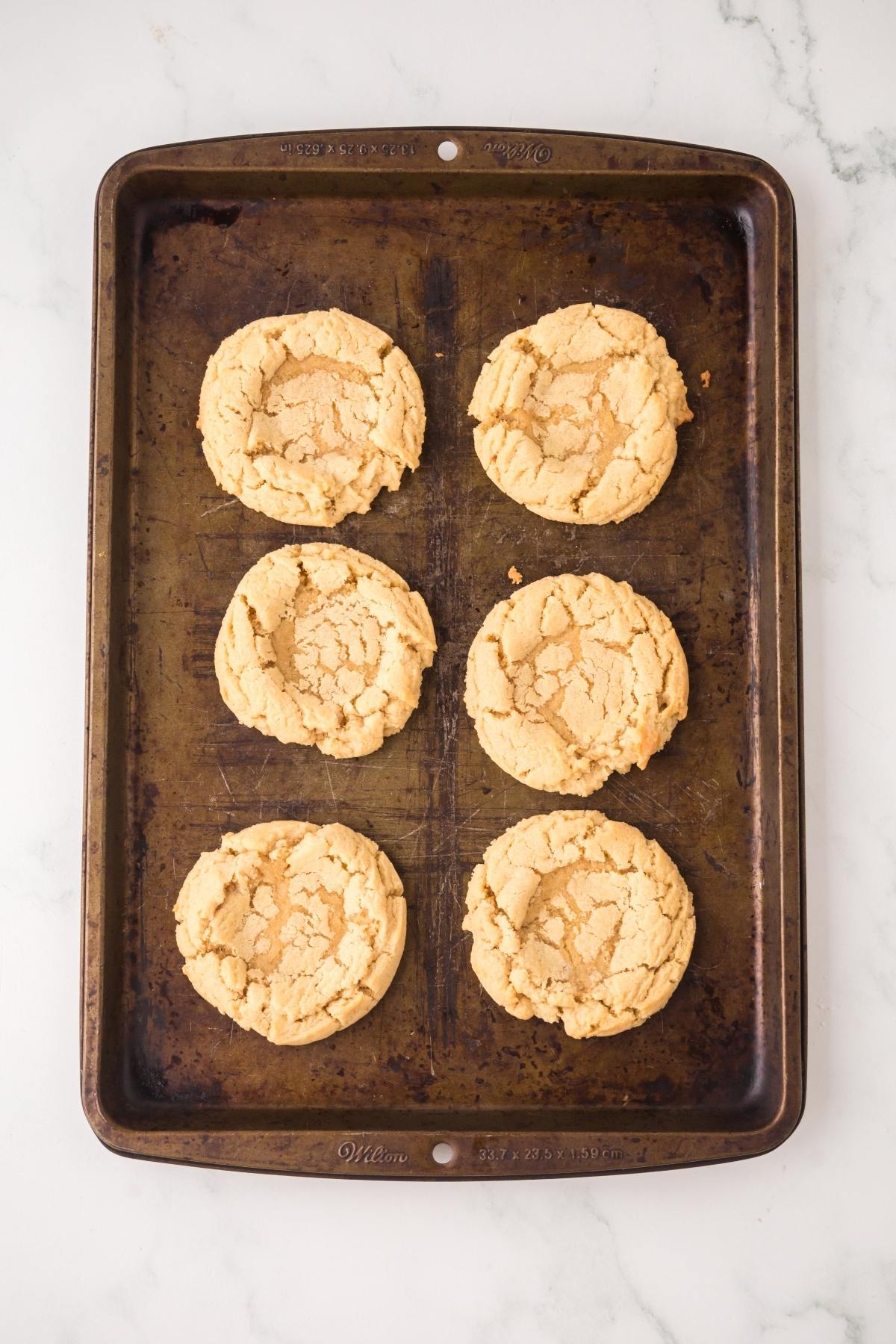 Six round, golden cookies on a dark metal baking sheet, set on a white marble surface.