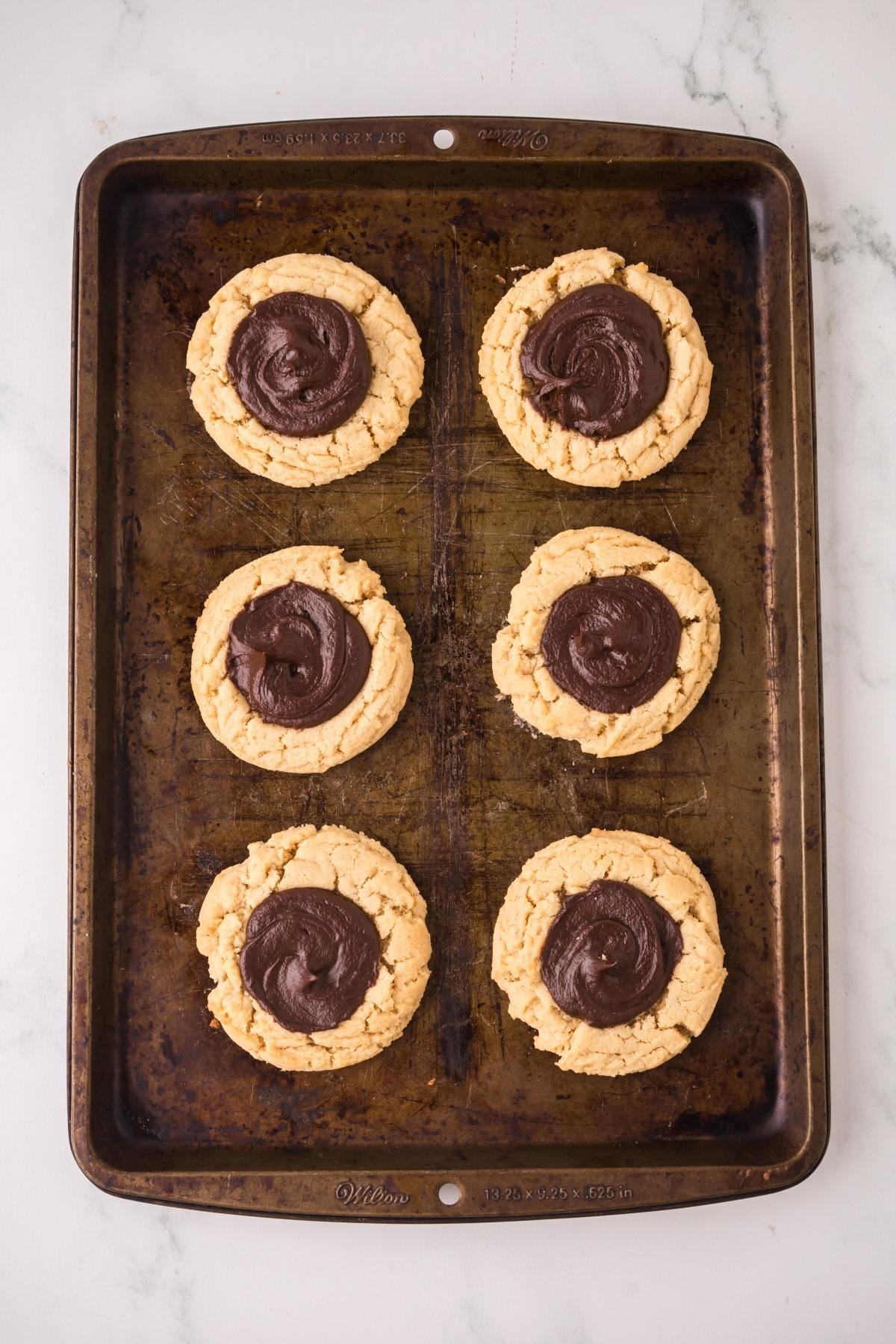 Six cookies with chocolate centers on a worn baking sheet, viewed from above on a white surface.