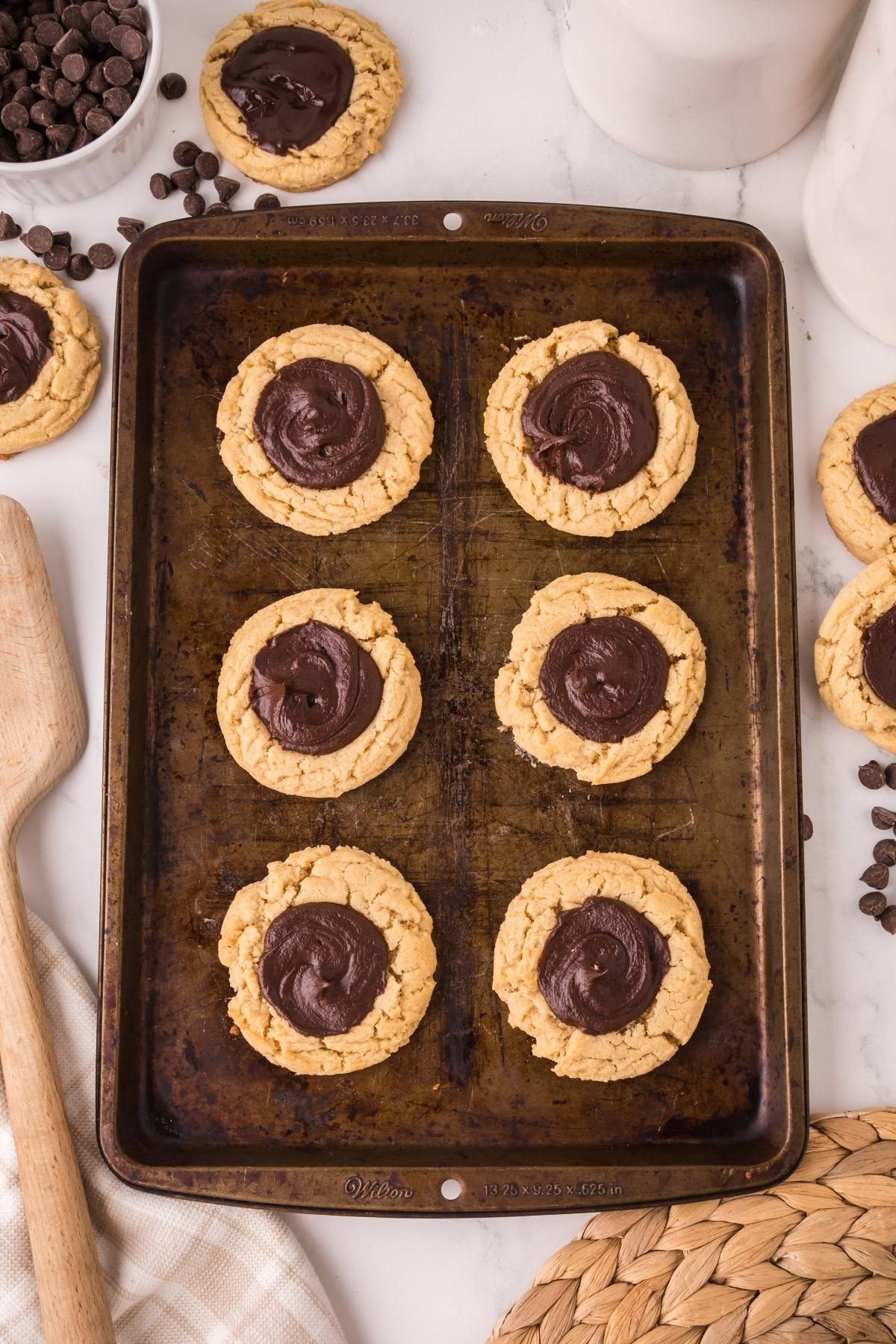 Eight chocolate thumbprint cookies on a baking sheet, surrounded by chocolate chips and cookie tools.