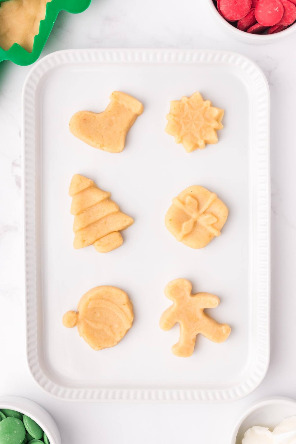 Six unbaked Christmas sugar cookies on a white tray in festive shapes like tree, stocking, and present.