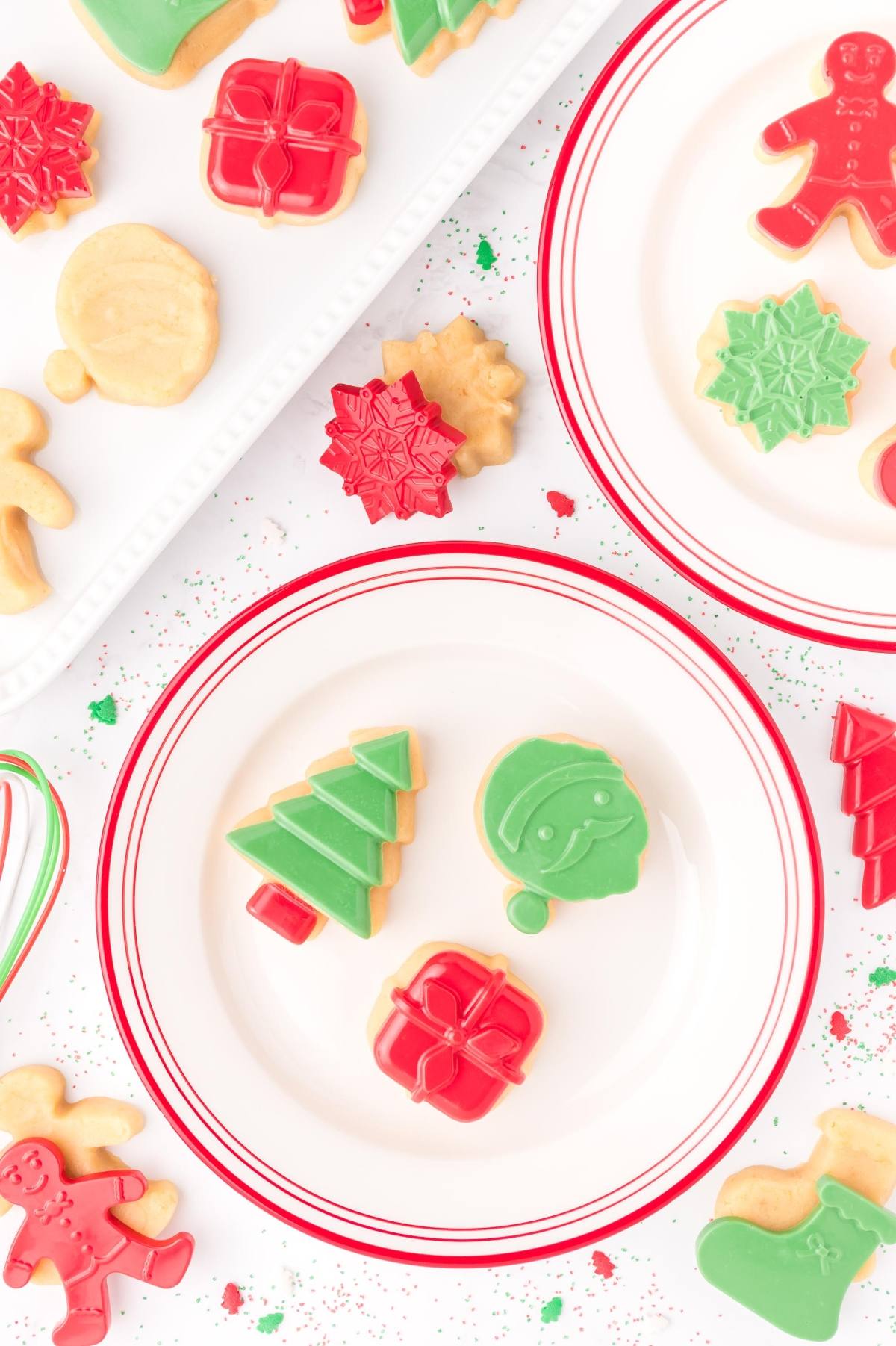 Christmas cookies shaped like trees, presents, snowflakes, and gingerbread men on white plates with red rims.