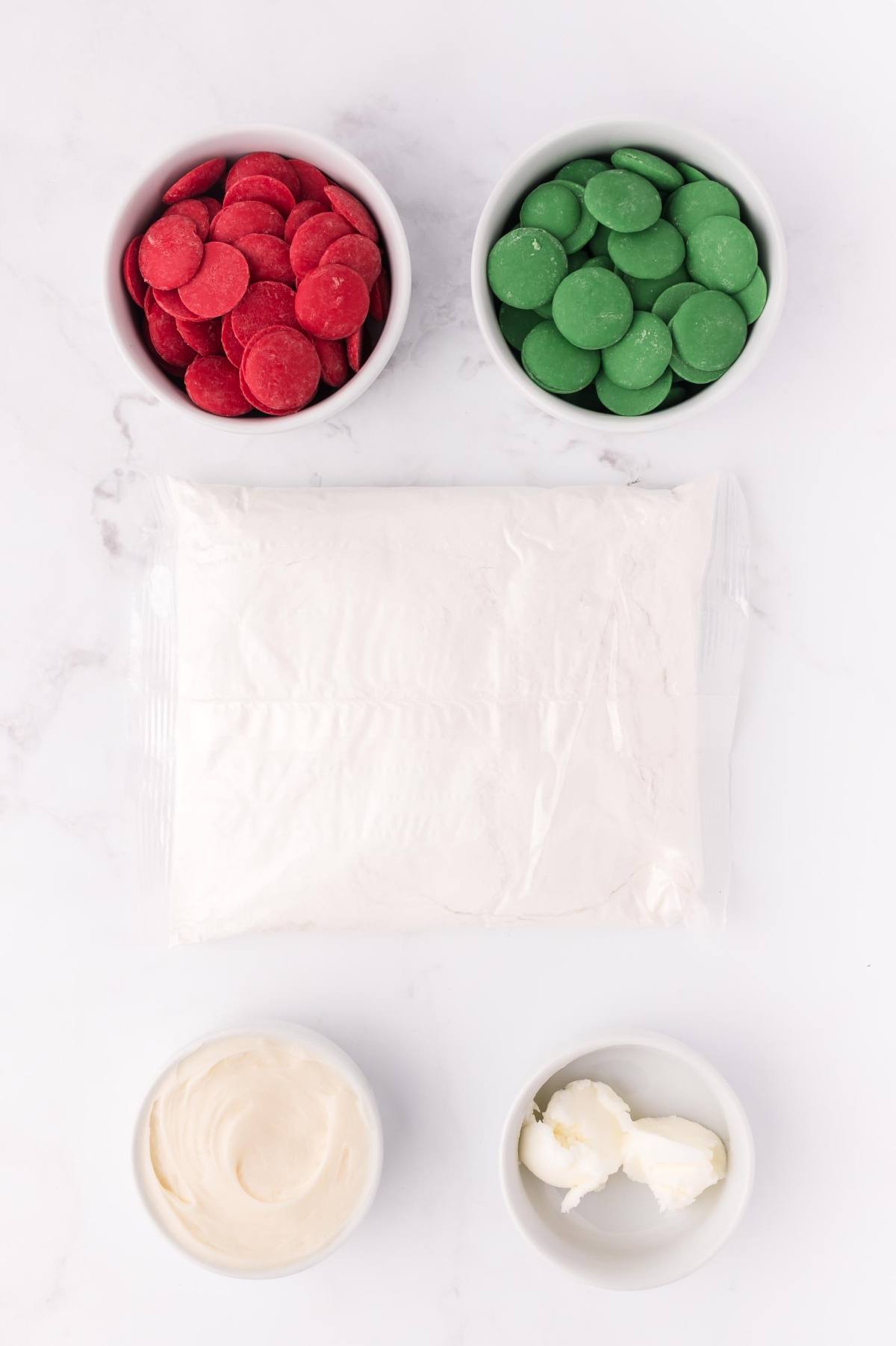 Red and green candy melts, a bag of white powder, white frosting, and shortening in small bowls on a white surface.
