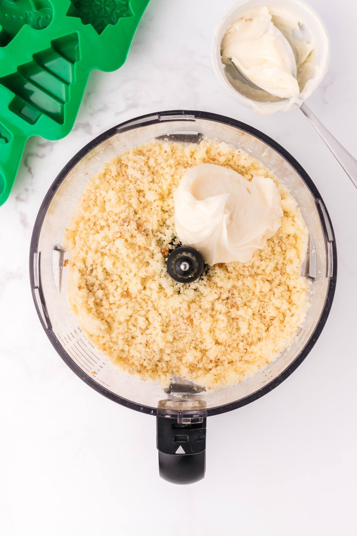 Food processor with cookie crumbs and cream cheese, beside a green tree mold and a bowl of cream cheese.