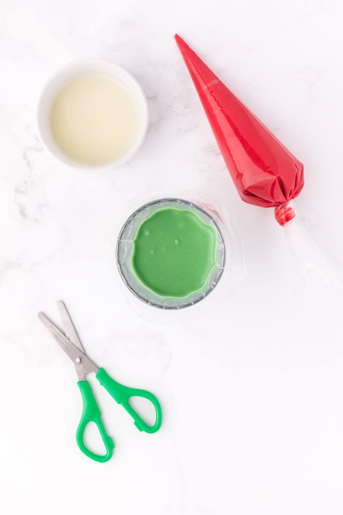 Green icing in a cup, red piping bag, scissors, and small bowl on a white surface.