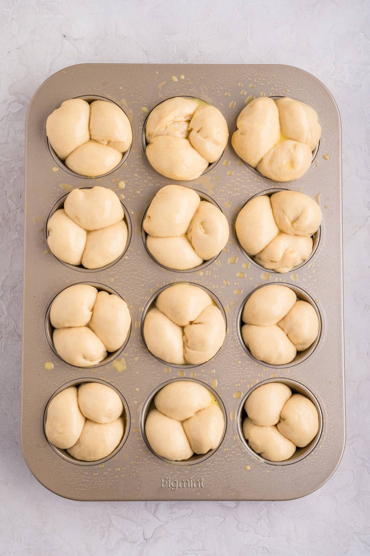 A muffin tin filled with unbaked, shaped bread rolls on a light surface.