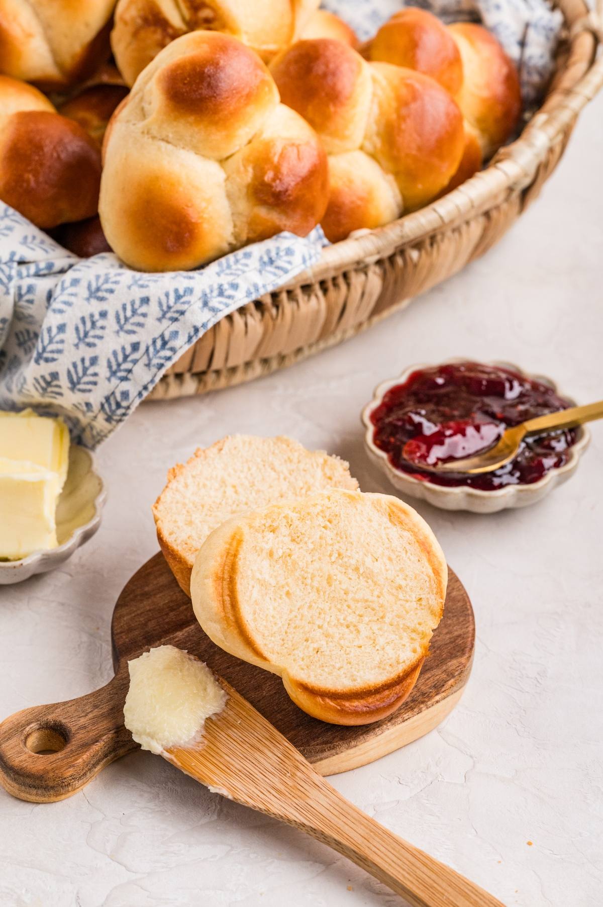 A sliced bread roll on a board with butter, jam, and a basket of more rolls in the background.
