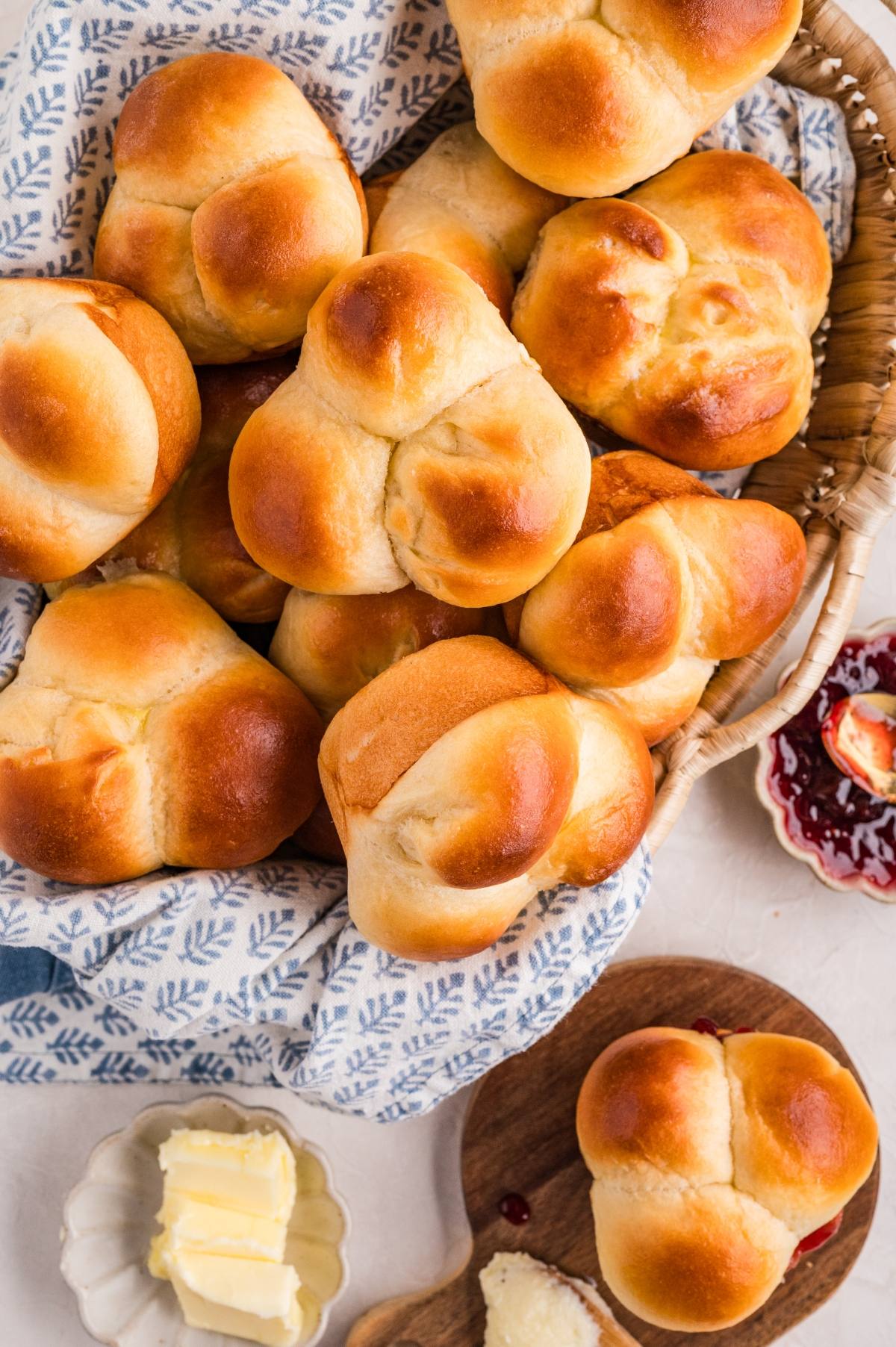 A basket of golden brown cloverleaf dinner rolls, with butter and jam on the side.
