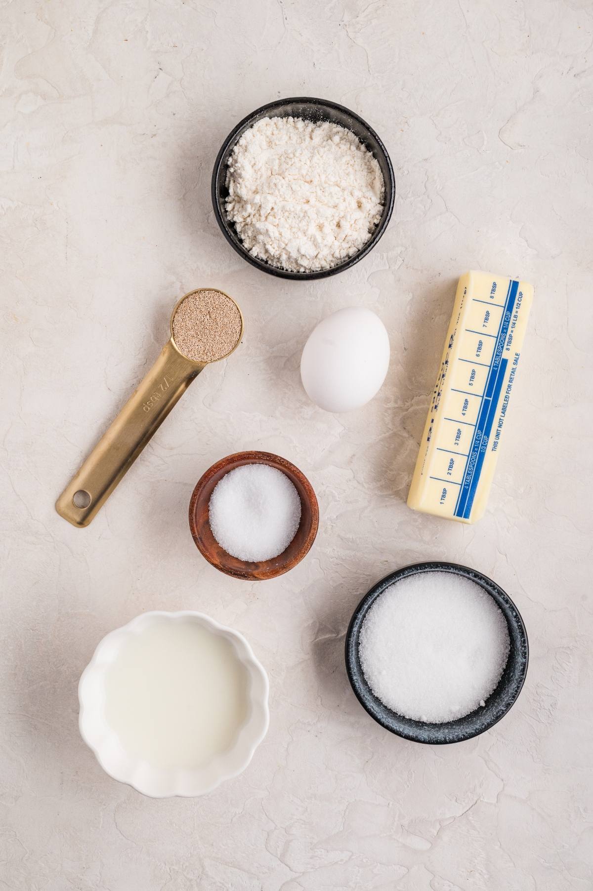 Baking ingredients: butter stick, egg, yeast, milk, salt, sugar, and flour in bowls on a white surface.