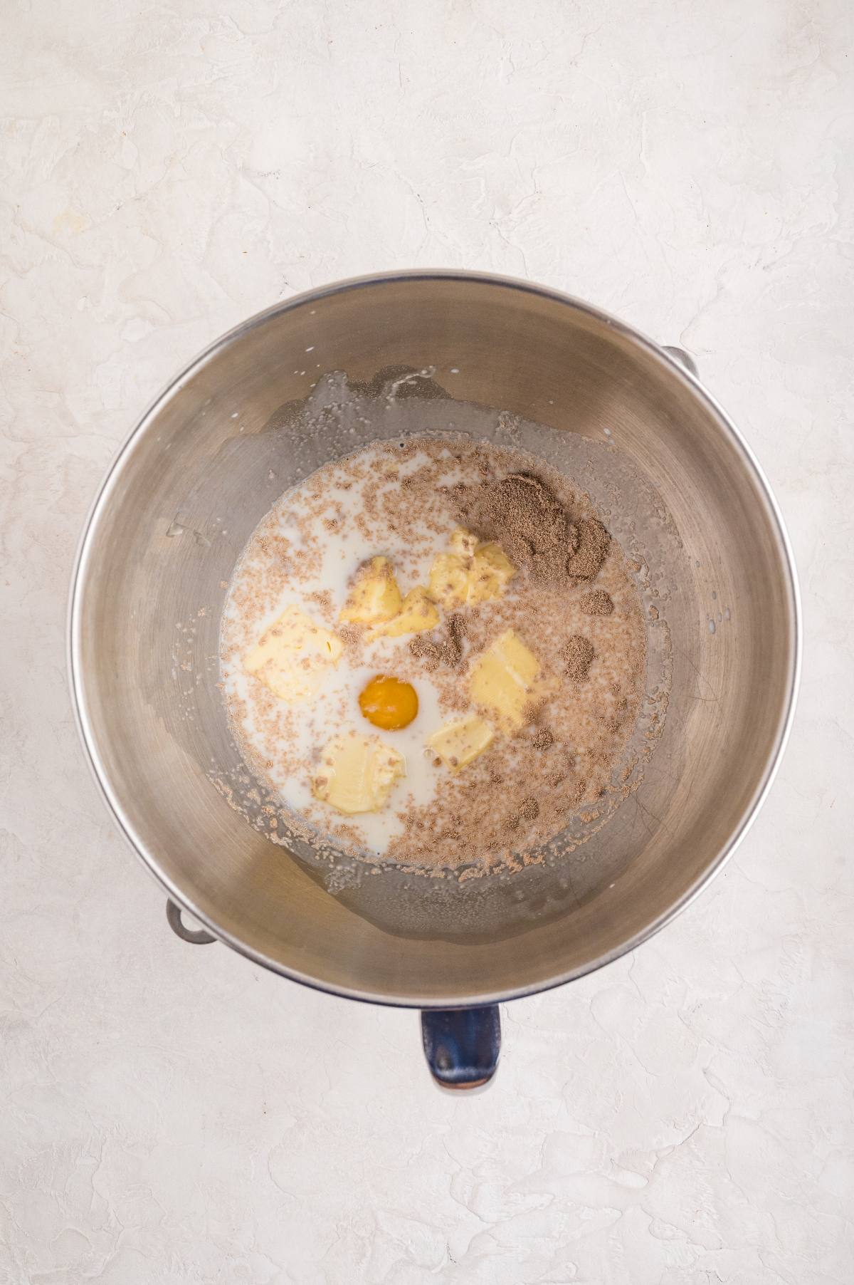 A mixing bowl with butter, an egg, brown sugar, oats, and milk on a light countertop.