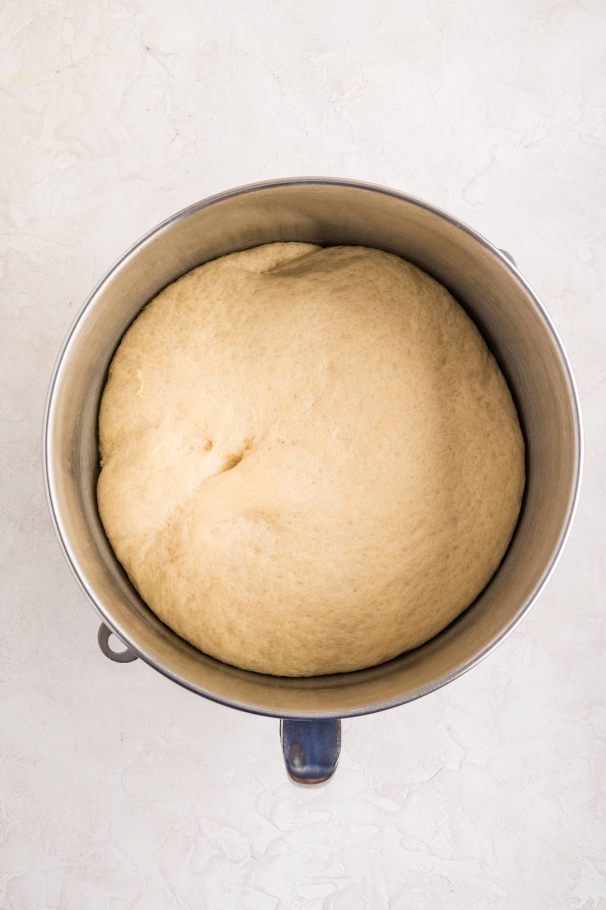 A bowl of risen bread dough in a metal mixing bowl on a light-colored surface.