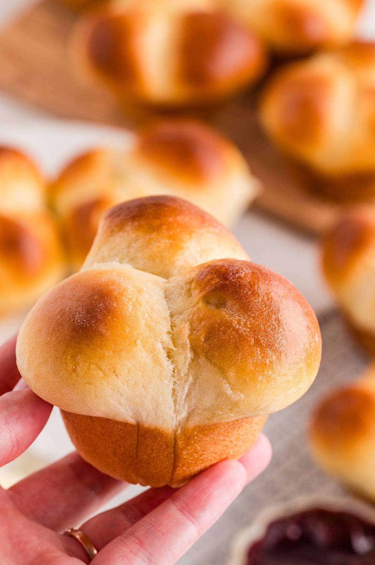 A hand holding a golden-brown cloverleaf dinner roll with more rolls in the background.