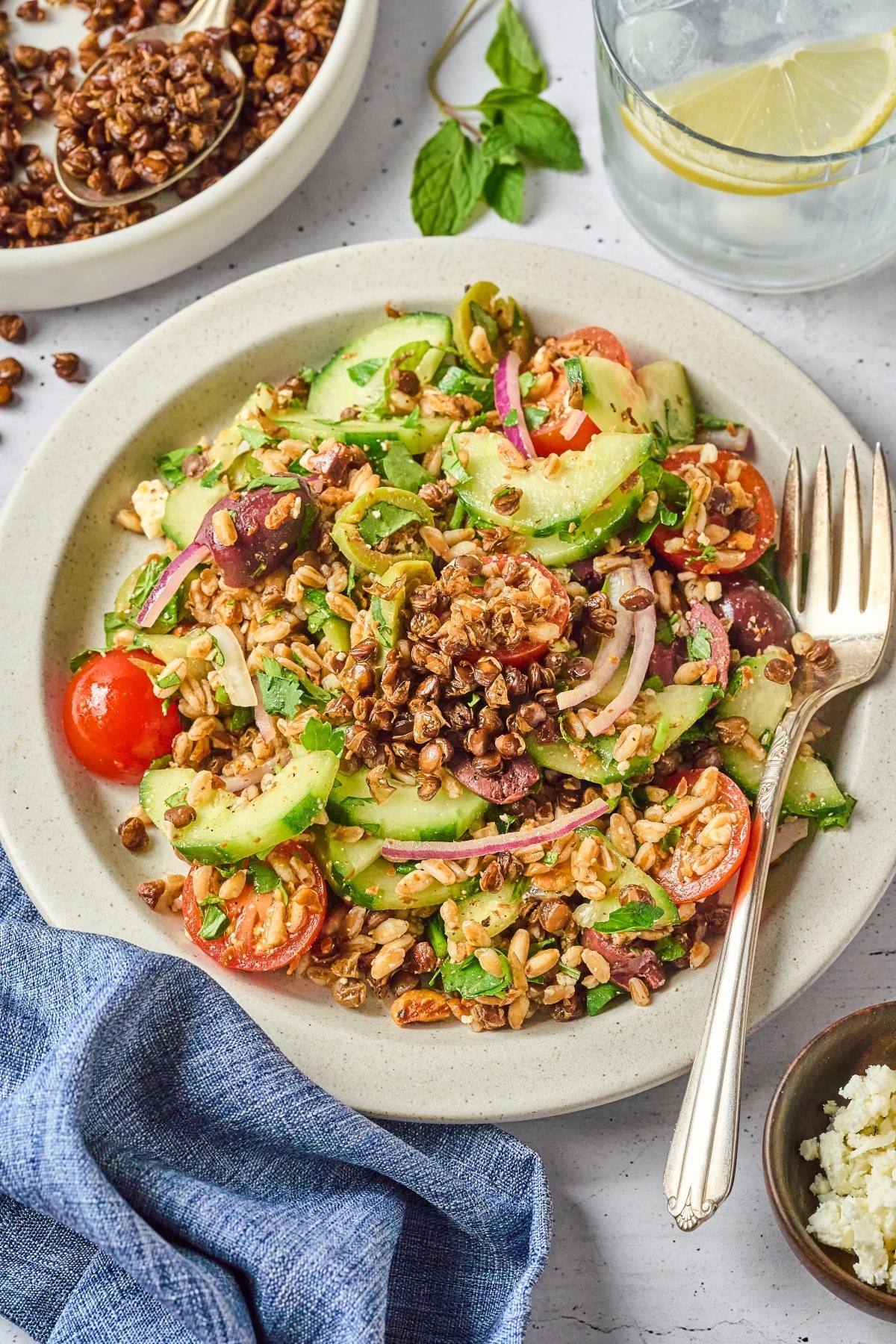 A plate of Greek salad with cucumbers, tomatoes, olives, grains, and crunchy toppings, next to a fork.