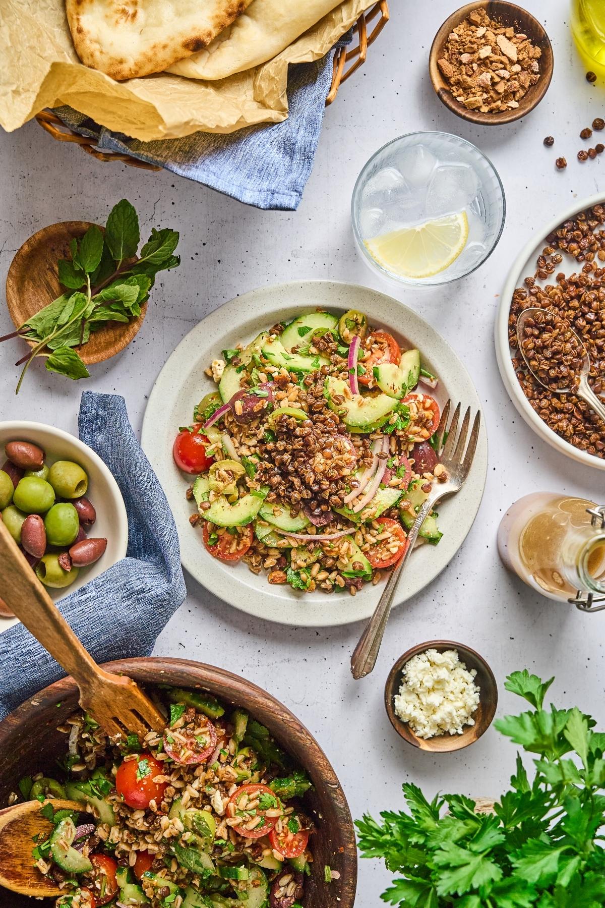 Colorful lentil salad with cucumbers, tomatoes, and feta on a plate, surrounded by bread, olives, and herbs.