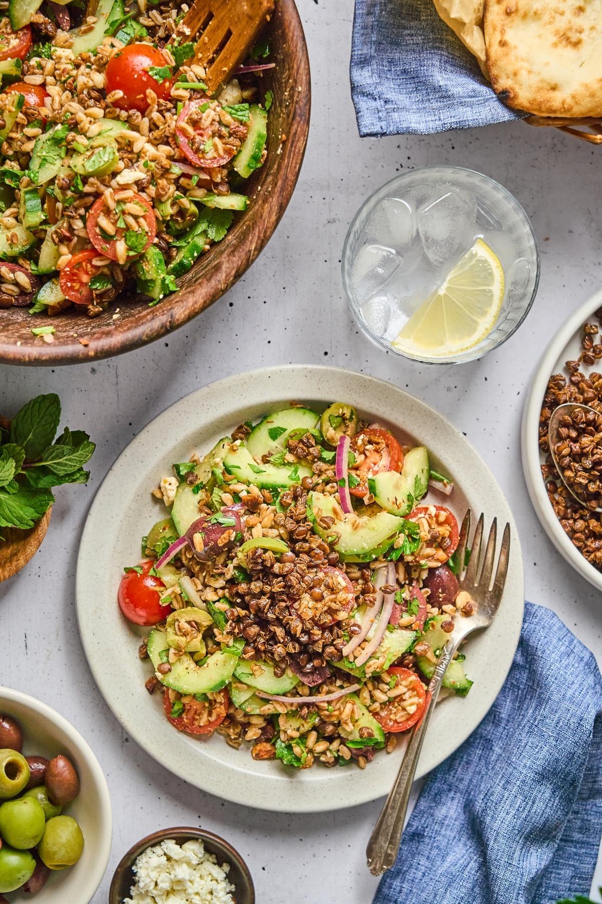 A plate of Greek salad with tomatoes, cucumber, olives, grains, and a glass of water with lemon.