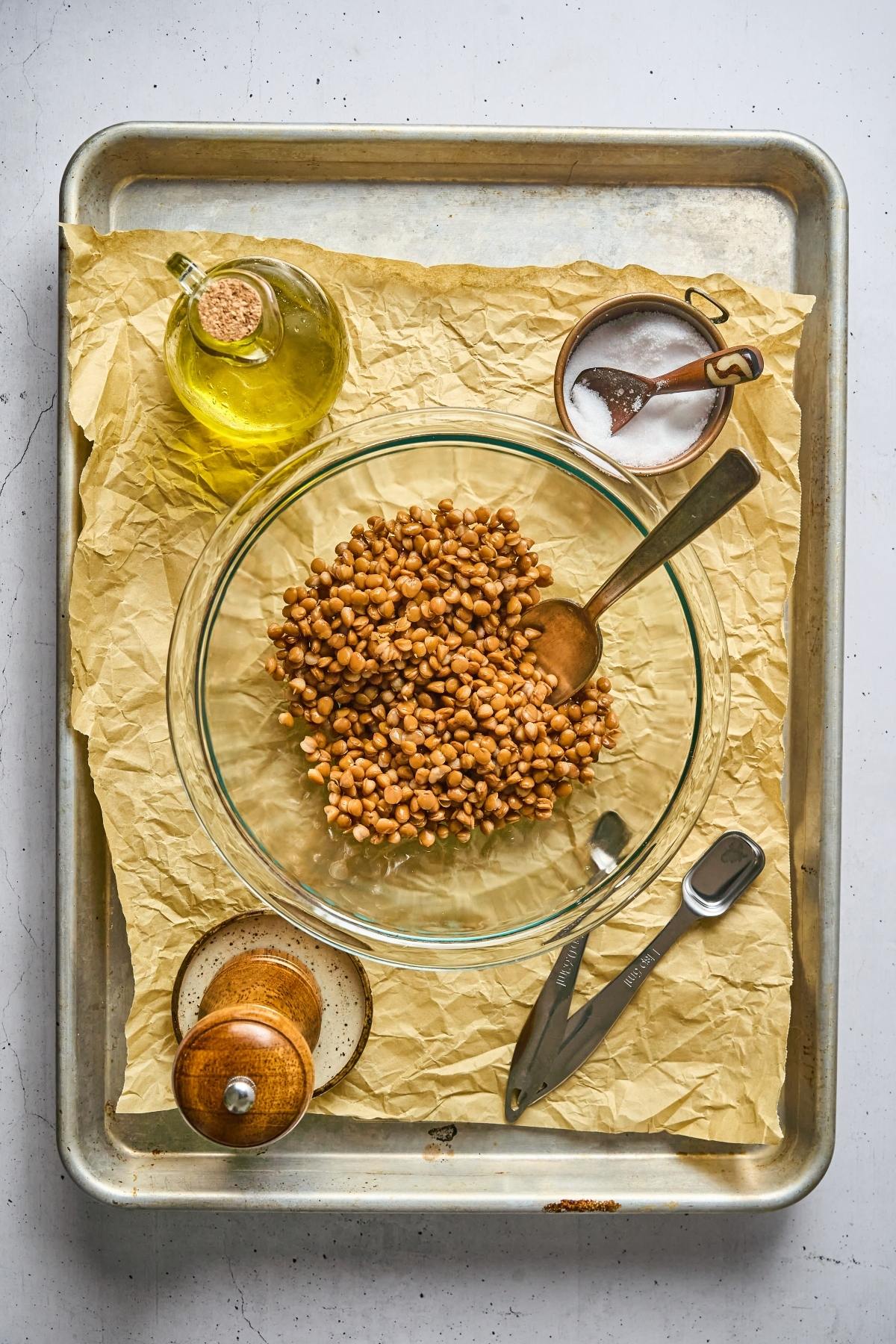 Glass bowl of chickpeas with spoon, olive oil, salt, pepper grinder, and measuring spoons on parchment paper.