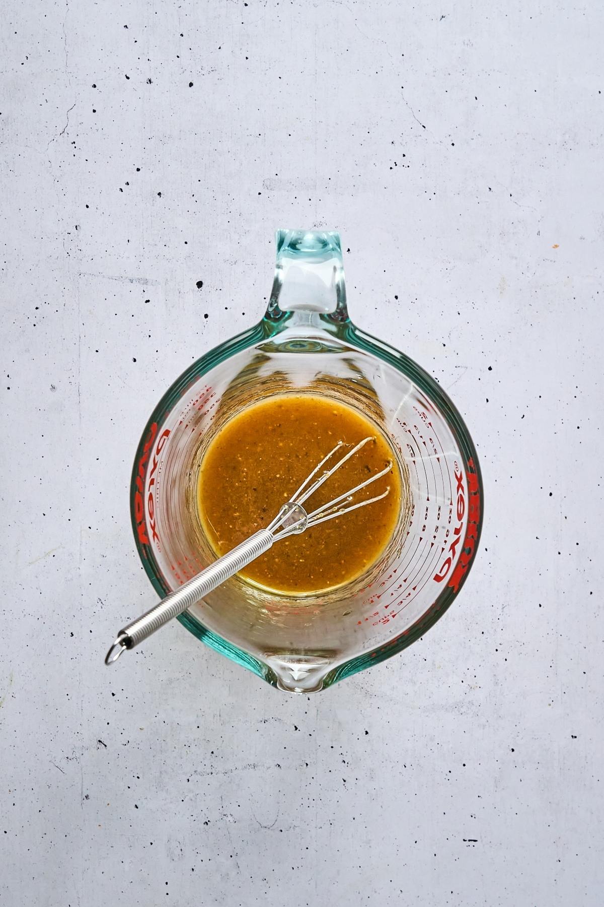 Glass measuring cup with brown liquid and a small whisk on a light, textured surface, viewed from above.