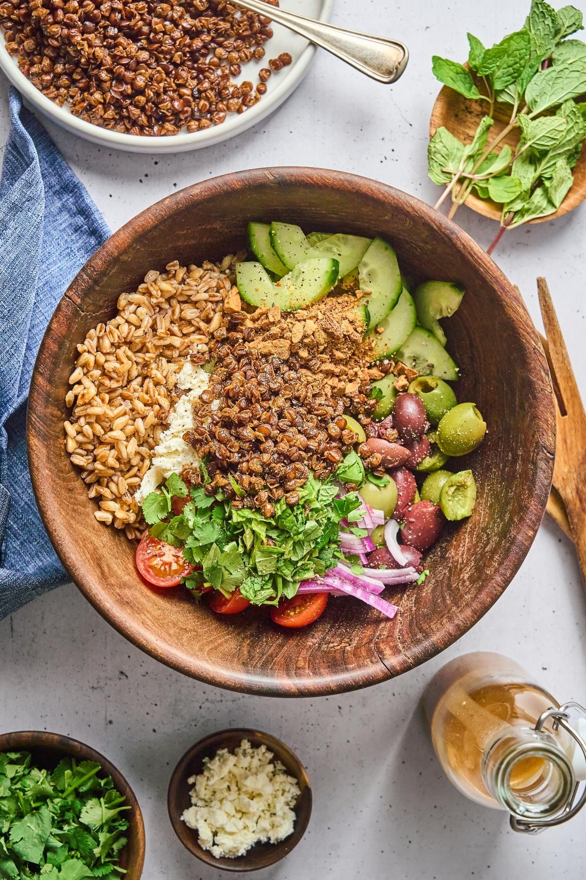 A wooden bowl with grains, lentils, cucumber, tomatoes, olives, onions, herbs, and feta cheese.