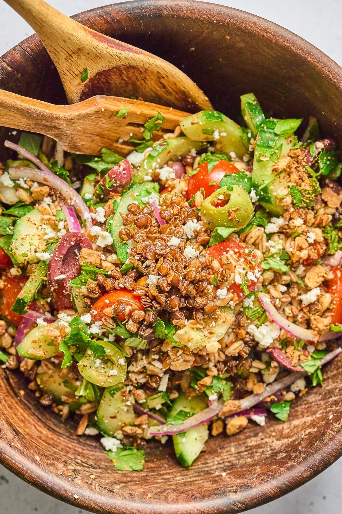 A wooden bowl of Greek salad with tomatoes, cucumbers, onions, feta, olives, seeds, and wooden utensils.