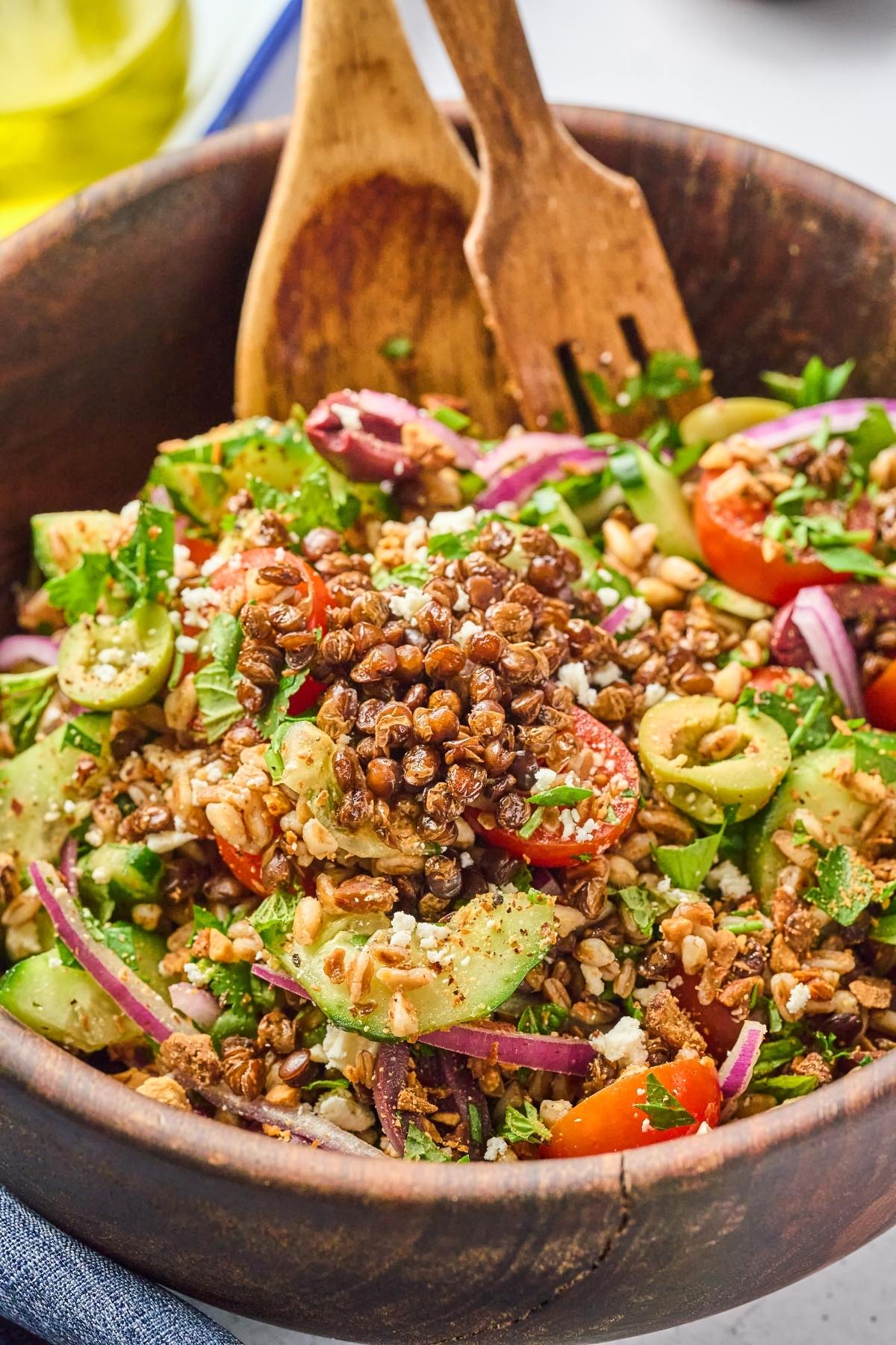 A wooden bowl of Mediterranean salad with lentils, cucumber, tomatoes, red onion, herbs, and wooden utensils.