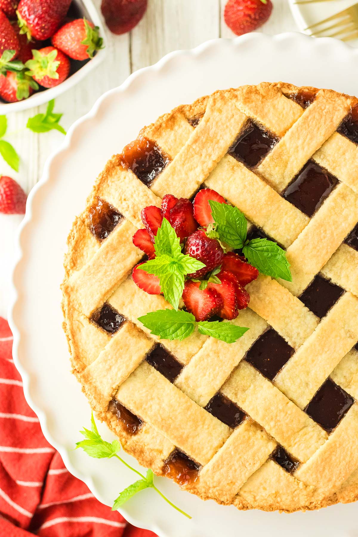 A lattice-top pie, crostata di mermellata, inspired by Italian dessert recipes, garnished with fresh strawberries and mint leaves, with extra strawberries on the side.