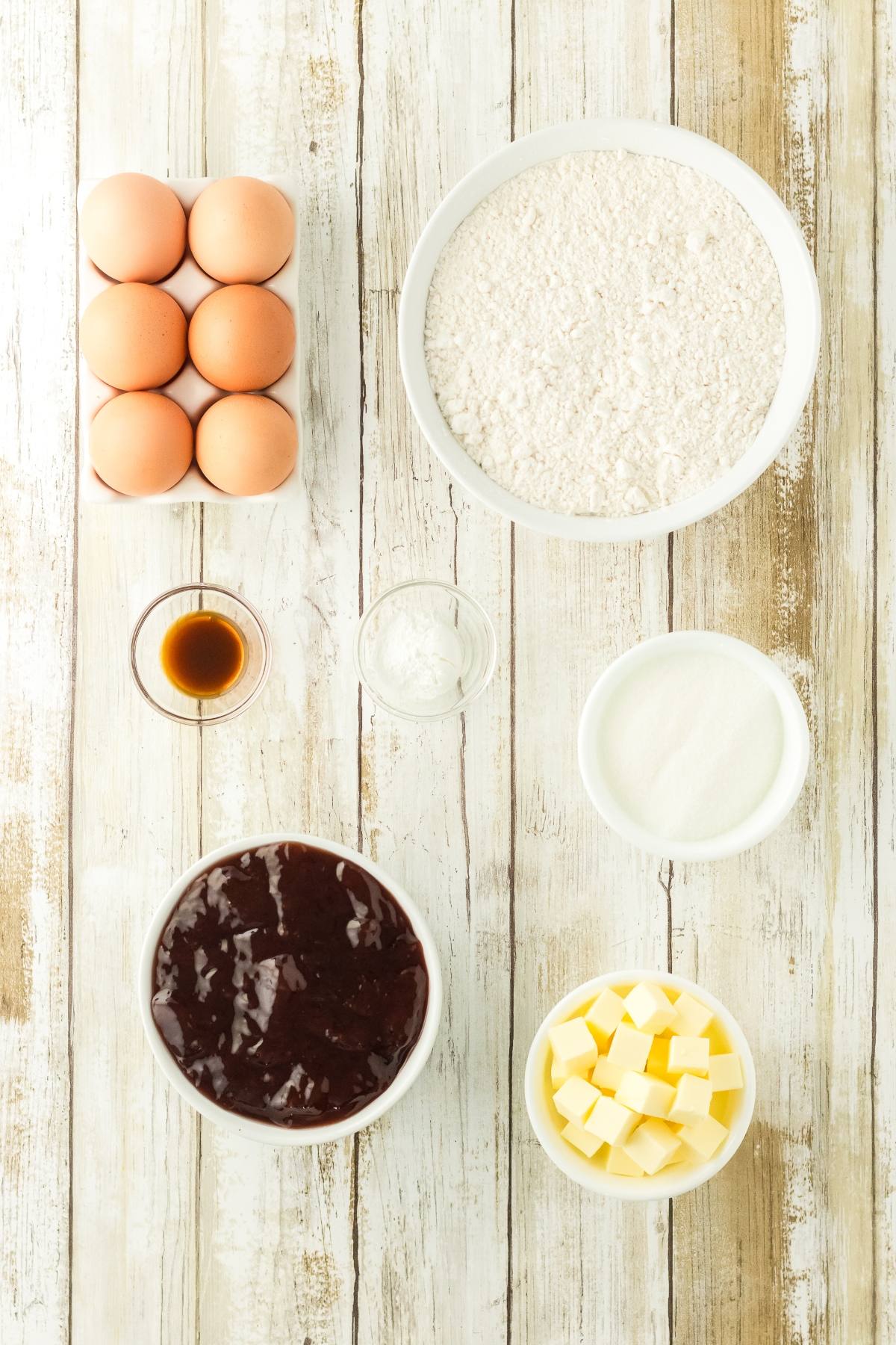 Baking ingredients on a wooden table: eggs, flour, vanilla, baking powder, sugar, butter, and chocolate batter.