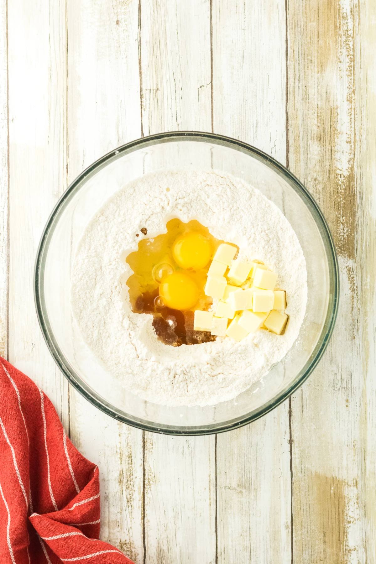 Glass bowl with flour, eggs, butter cubes, and vanilla on a wooden surface, red striped towel beside it.