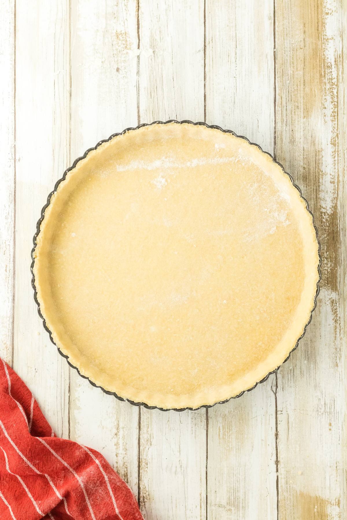 Unbaked pie crust in a round tart pan on a white wooden surface, with a red striped cloth nearby.