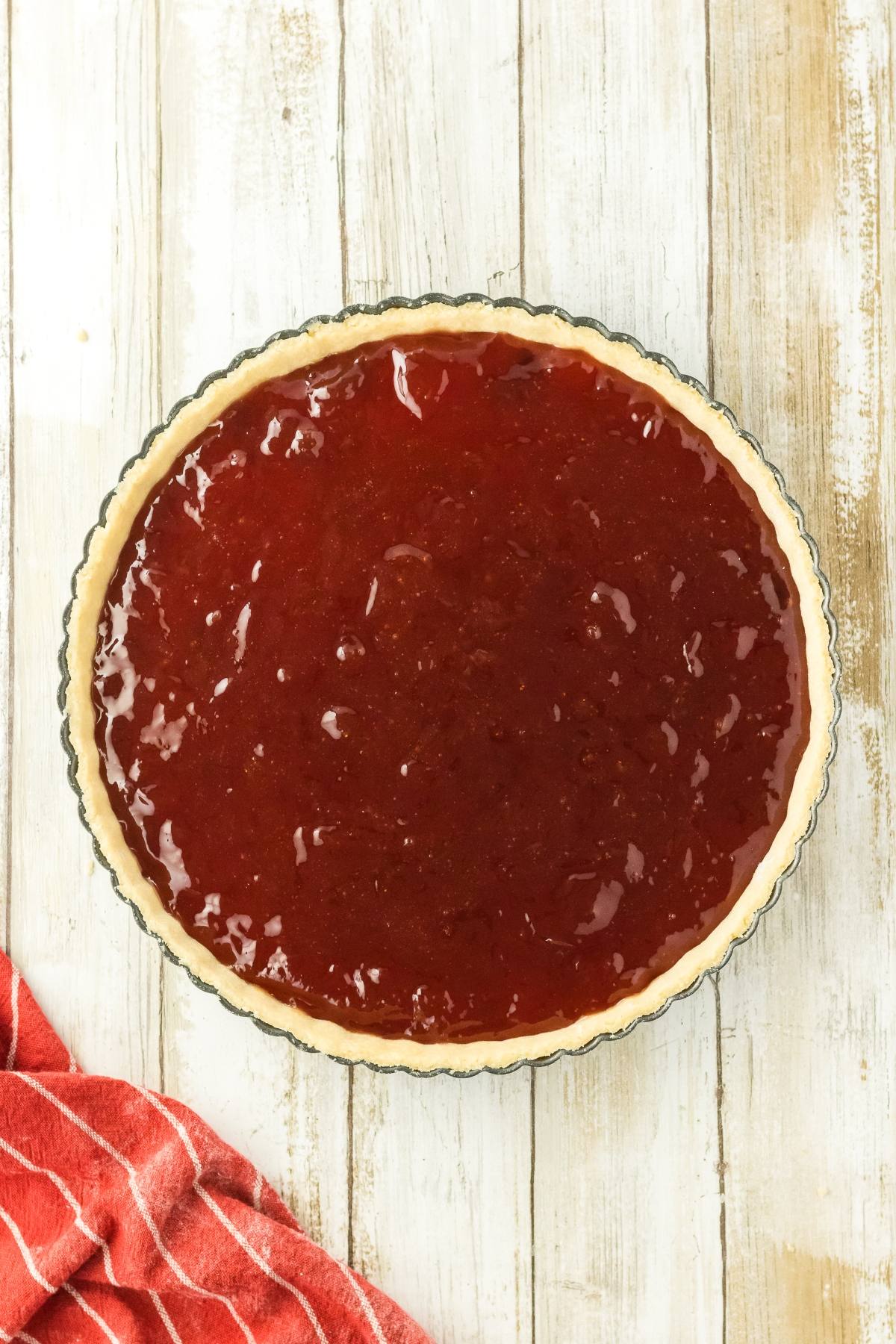 A tart crust filled with glossy red strawberry or raspberry jam on a wooden surface, next to a red towel.