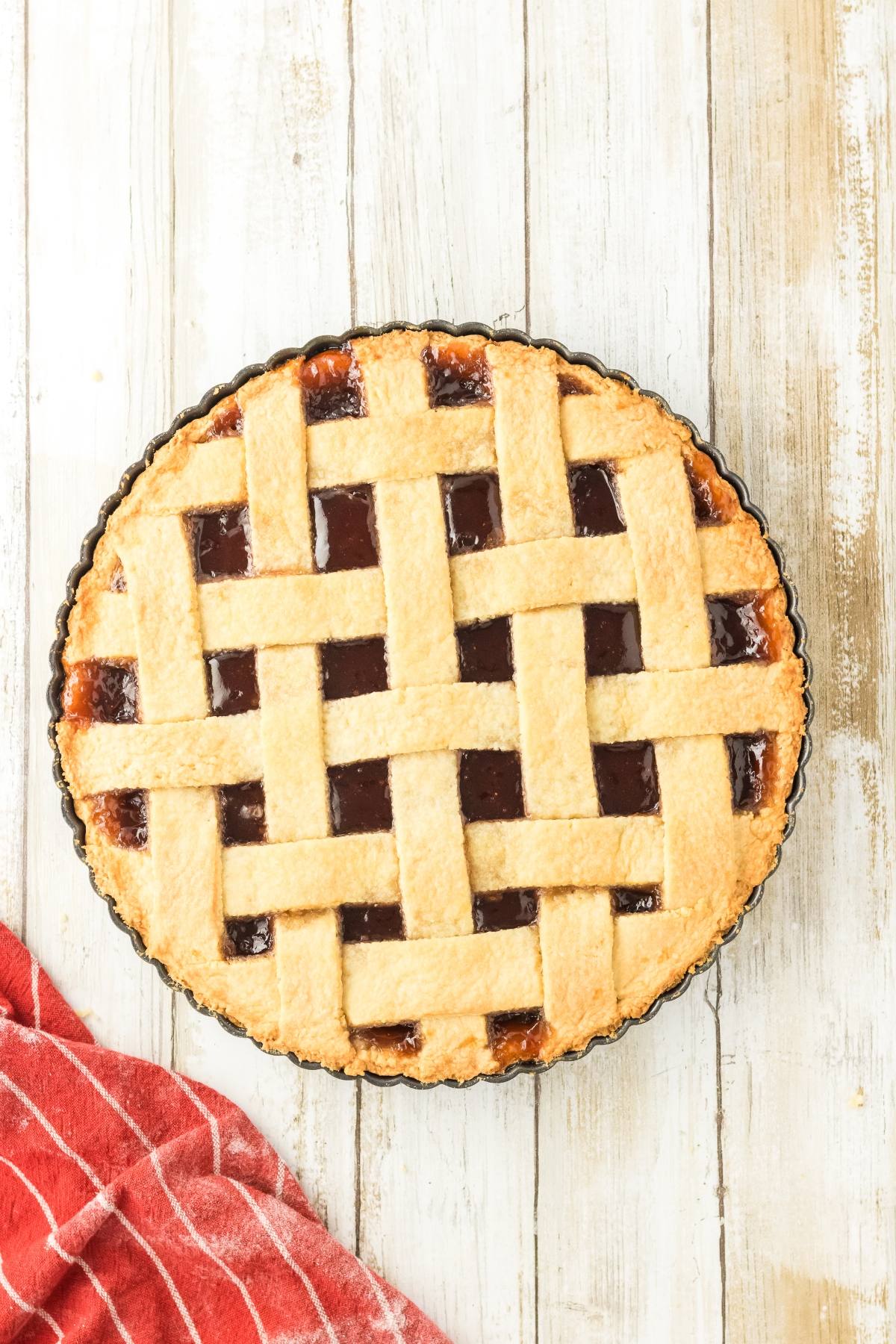 A cherry pie with a golden lattice crust sits on a white wooden table next to a red kitchen towel.