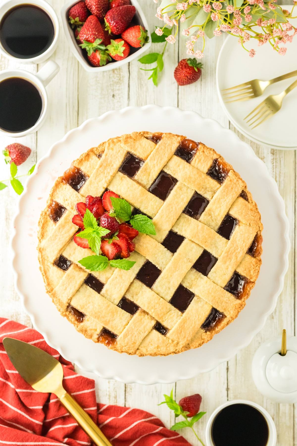 Lattice-top berry pie on a white plate, with coffee, strawberries, and gold utensils on a white table.