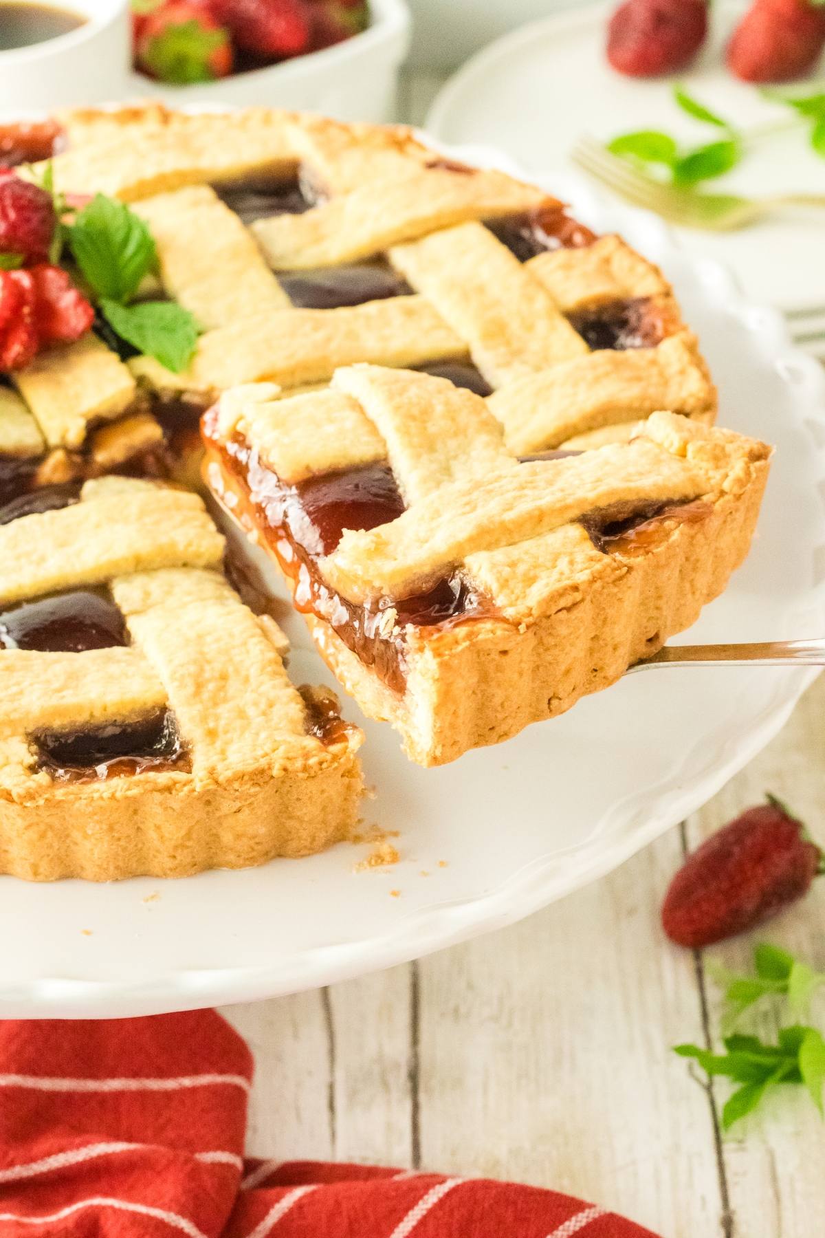 A lattice-topped fruit pie with a slice being lifted, on a white cake stand.