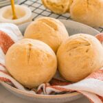 Three crusty rolls sit in a bowl lined with a red and white striped towel, with more rolls visible in the background.