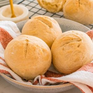 Three crusty rolls sit in a bowl lined with a red and white striped towel, with more rolls visible in the background.