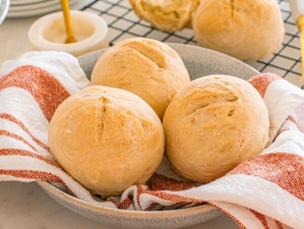 Three crusty rolls sit in a bowl lined with a red and white striped towel, with more rolls visible in the background.