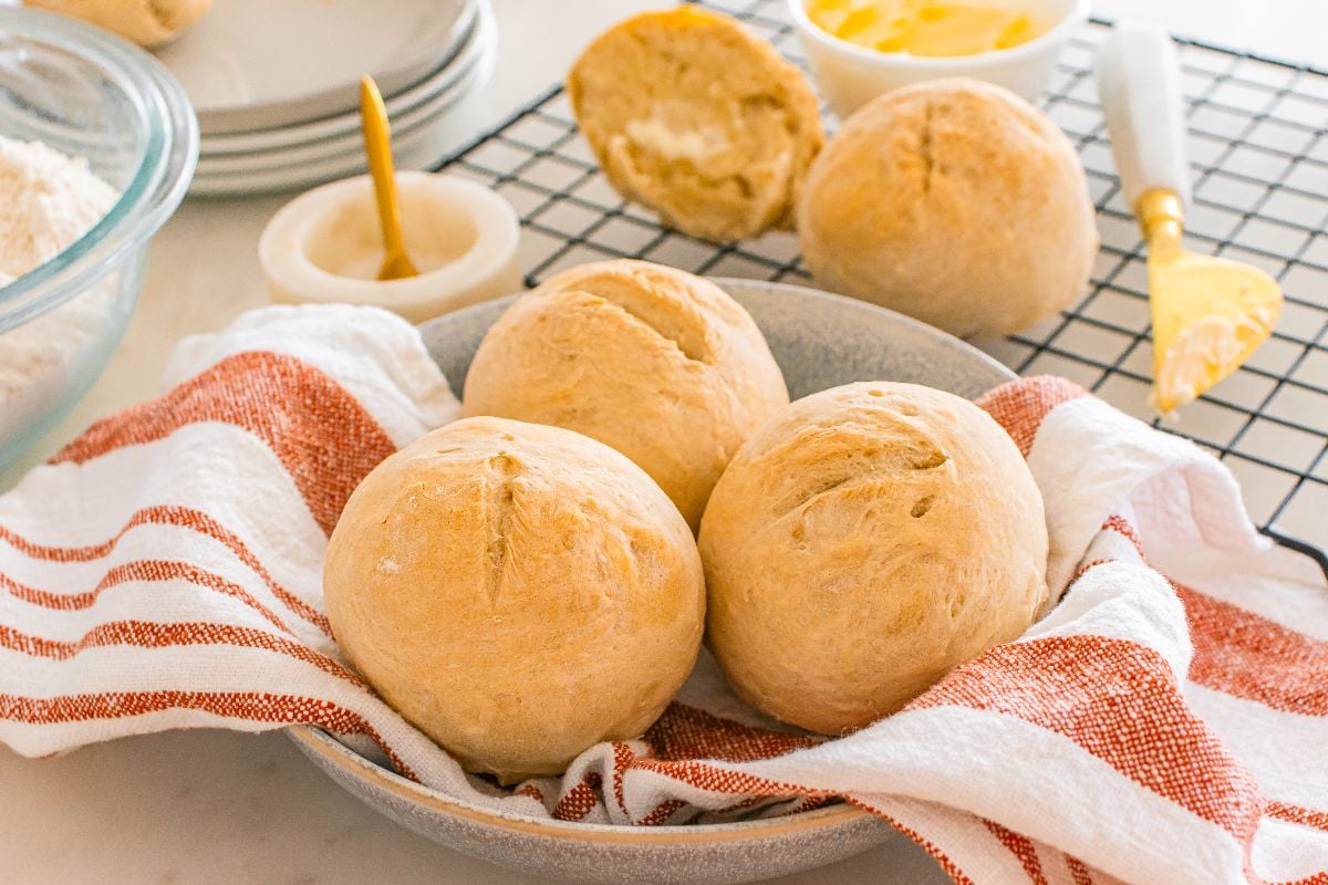 Three crusty rolls sit in a bowl lined with a red-striped towel, with butter and more golden rolls in the background.