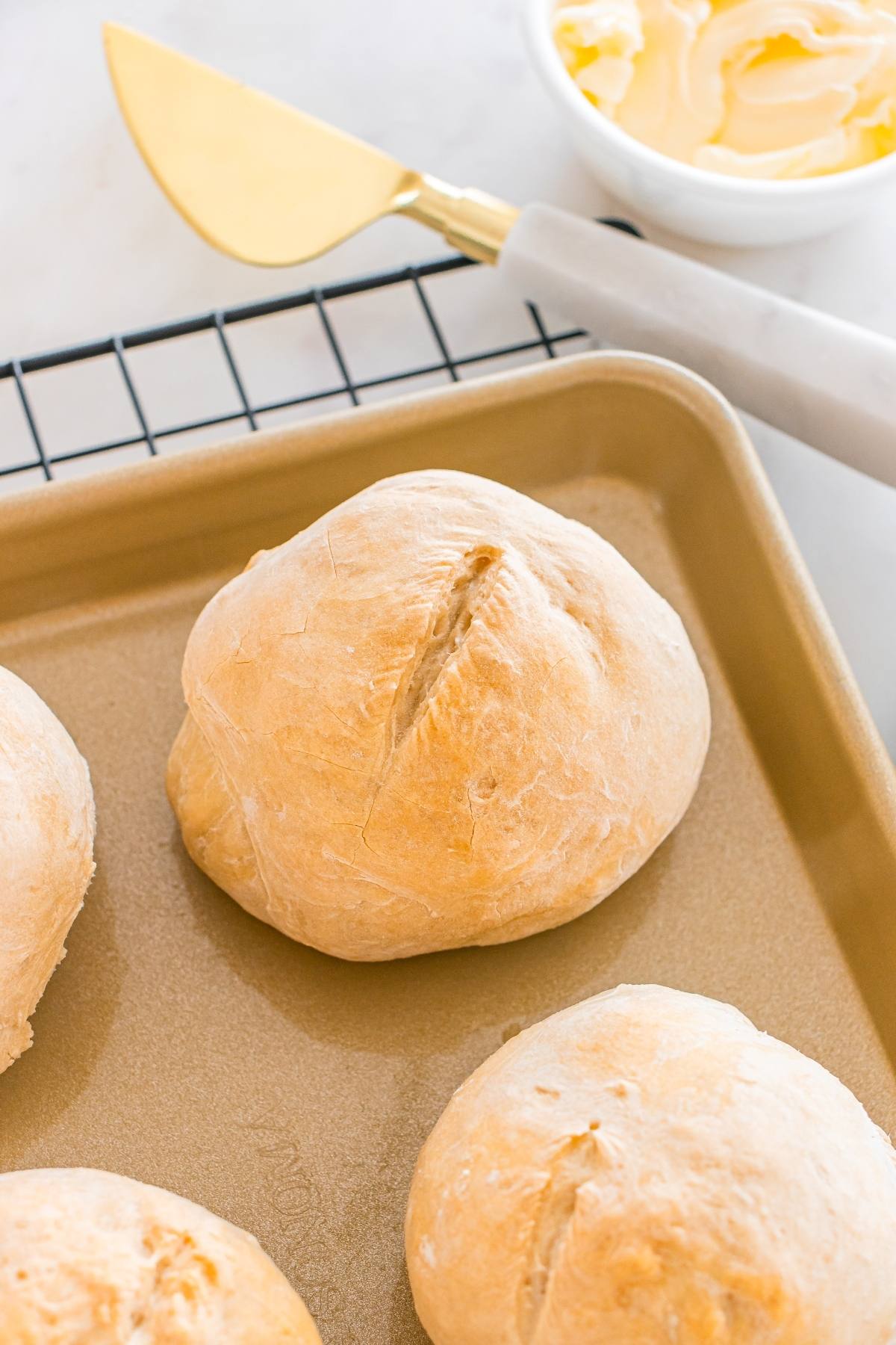 A golden brown bread roll on a baking sheet, with a bowl of butter and butter knife nearby.
